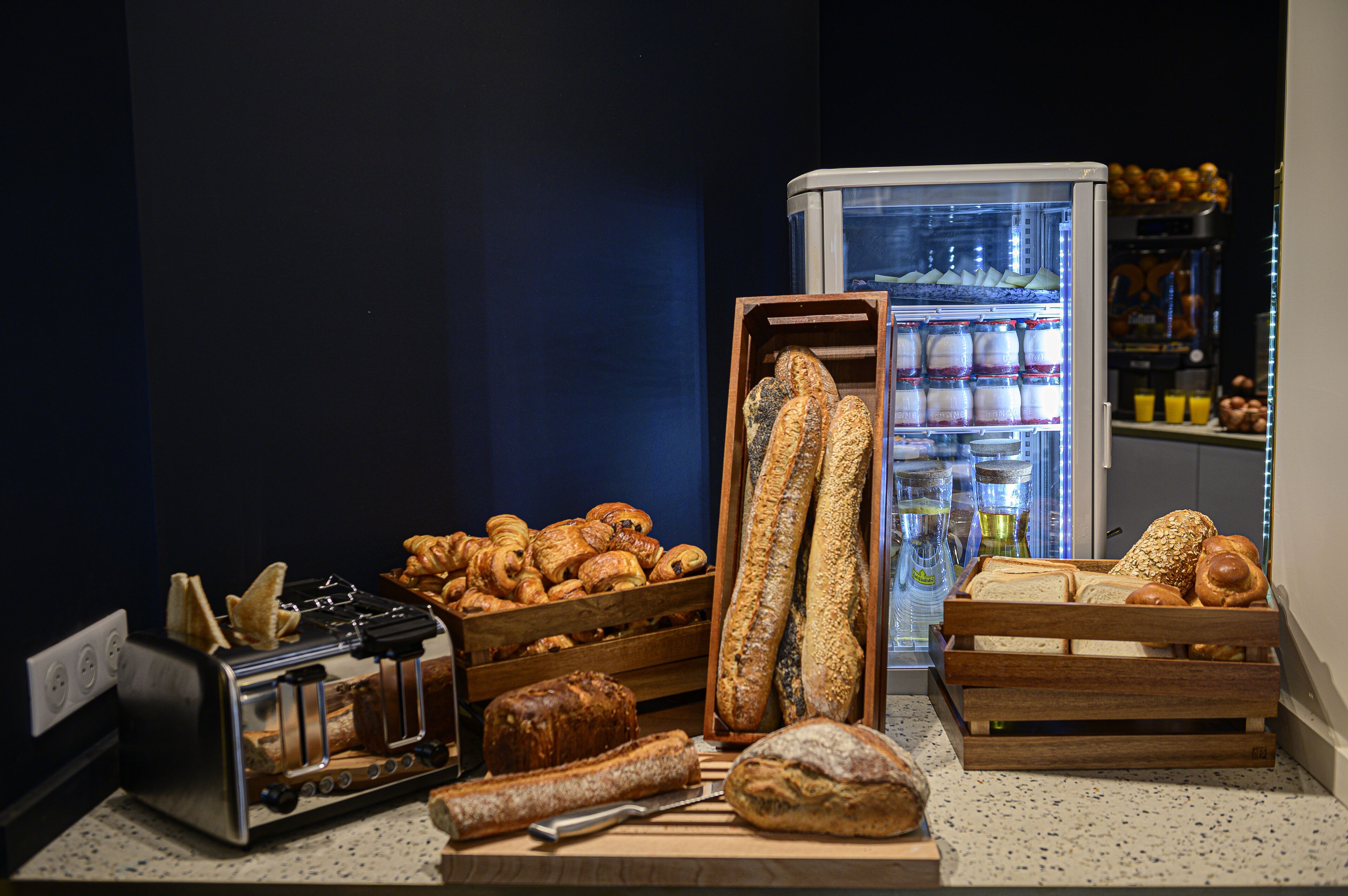 A bakery display with various breads and pastries, a toaster, and a refrigerator in the background.
