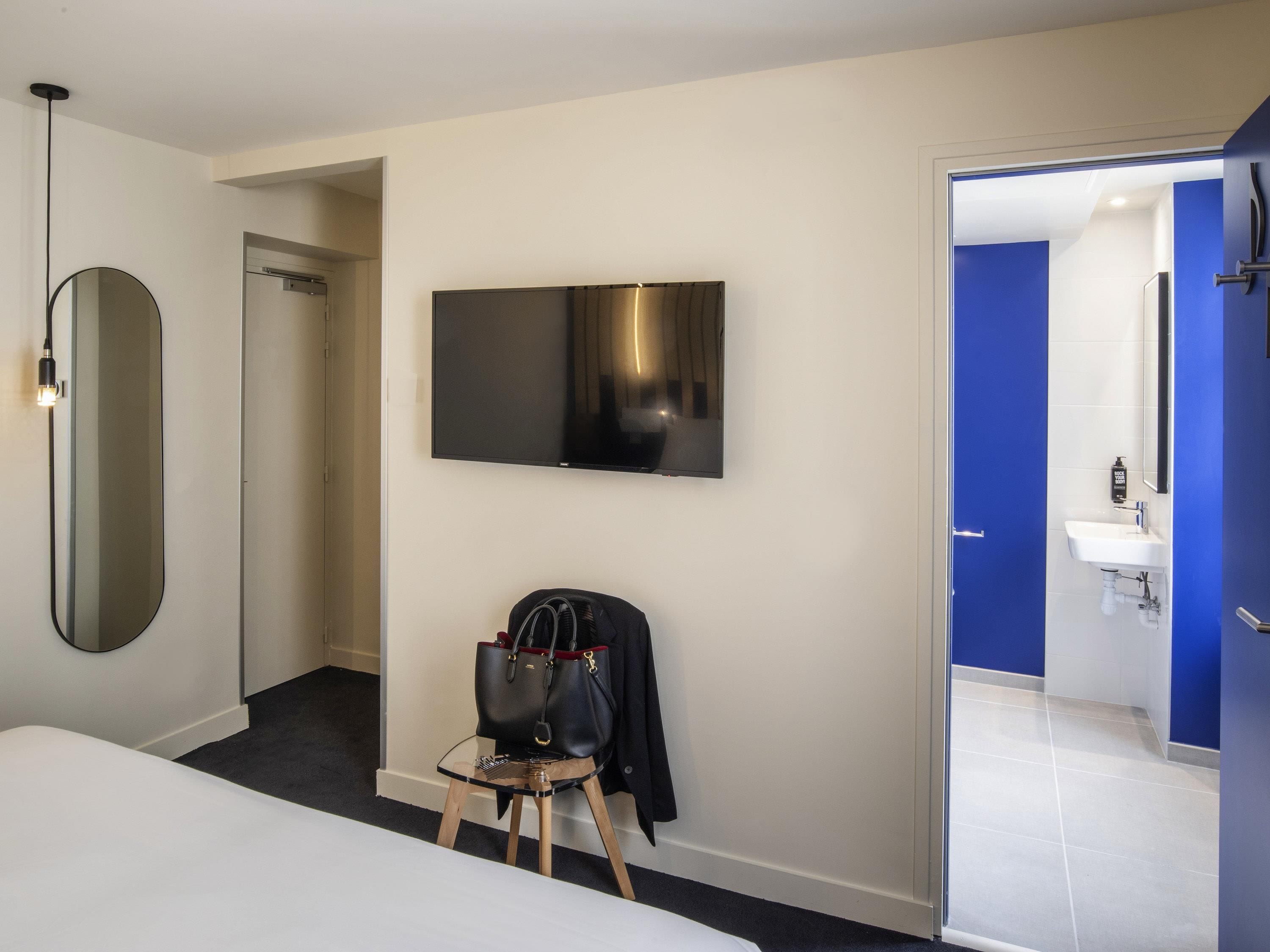 Una habitación de hotel moderna con una cama, un televisor instalado en la pared, un espejo, un taburete pequeño con una bolsa, y un baño visible a través de una puerta abierta.