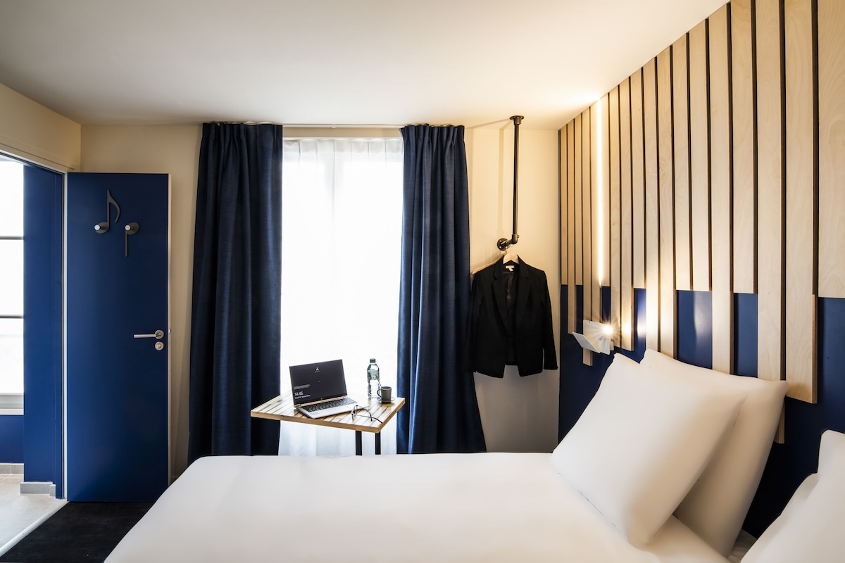 A hotel room with a bed, a desk with a laptop, a hanging coat, and a window with curtains.