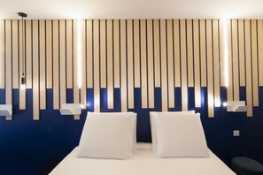 Habitación de hotel con cama, dos almohadas, mesita de noche y una lámpara fijada a la pared.