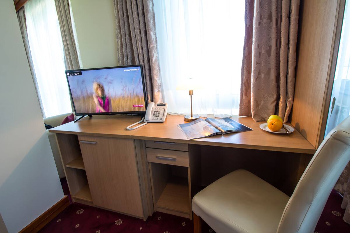 A hotel room with a desk, a telephone, a lamp, a plate with an orange, and a window with curtains.