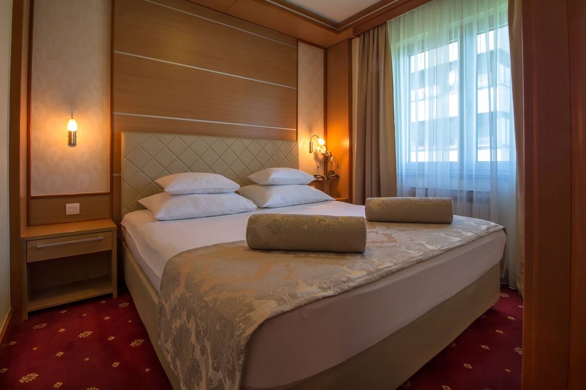 A hotel room with a large bed, two bedside tables, a nightstand, and a window with curtains.