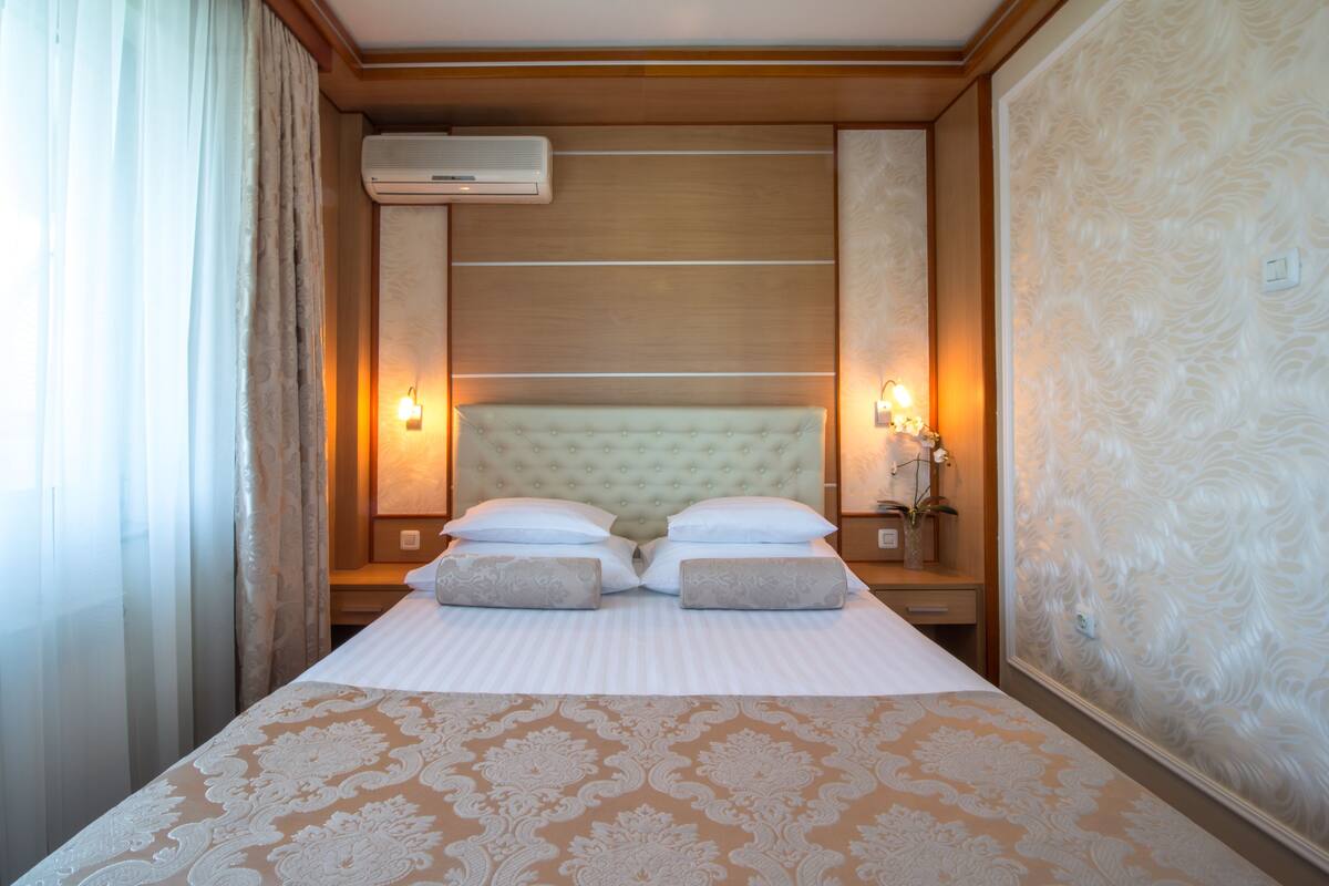 A hotel room with a large bed, two bedside lamps, an air conditioning unit, and a patterned wall.