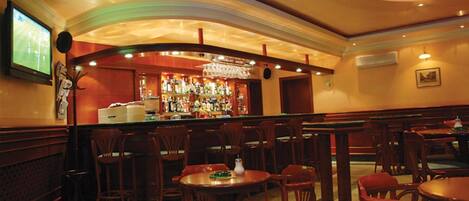 Uma área de bar com cadeiras e mesas de madeira, balcão de bar com garrafas e uma tela de televisão.
