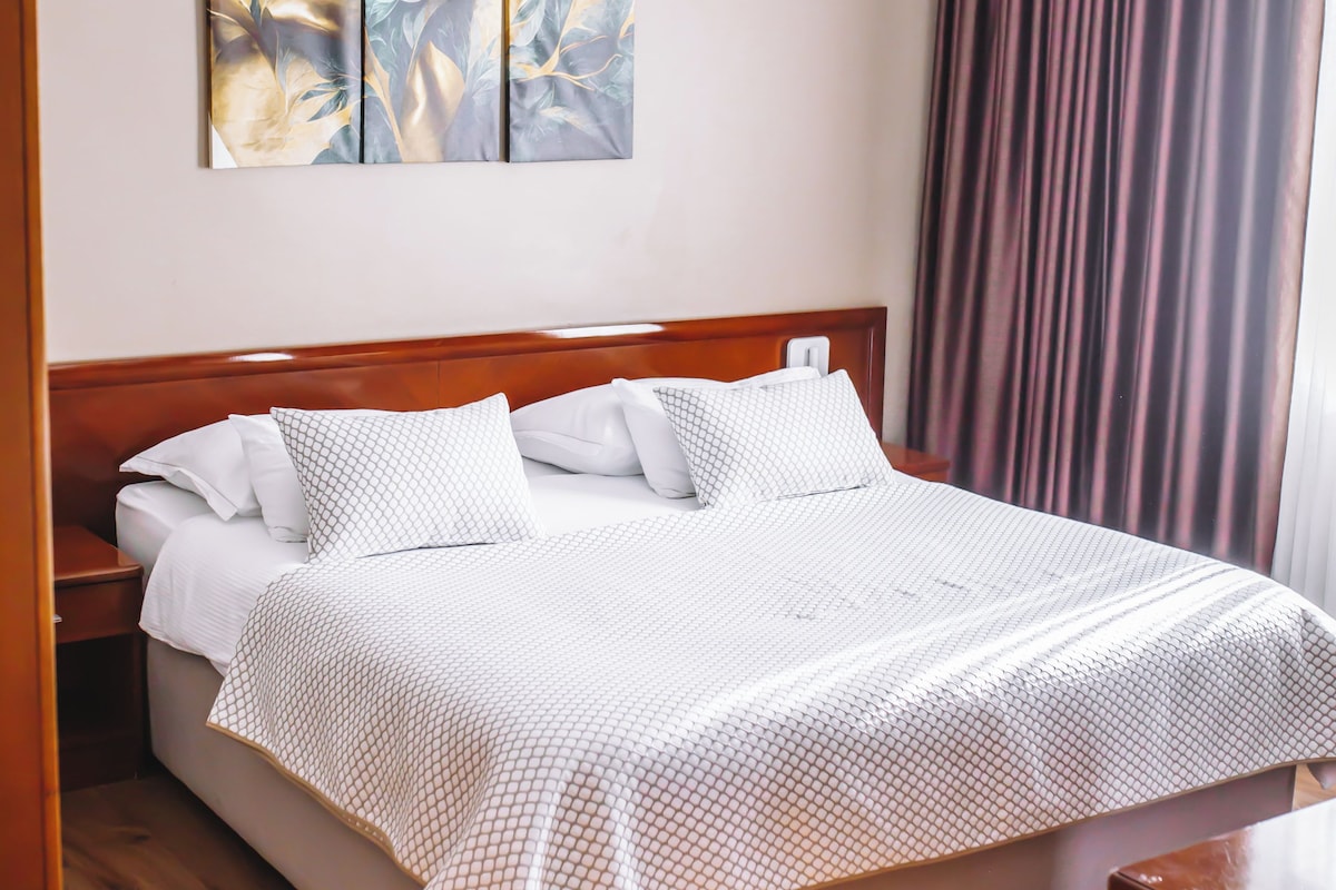 A neatly made bed with a white and patterned bedspread, a wooden headboard, and a wall-mounted artwork above.