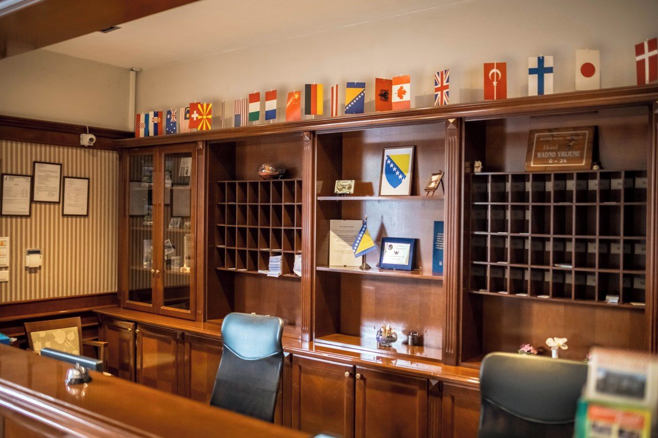 A formal office setting with a long wooden desk, two chairs, and shelves with flags and framed certificates.