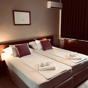 A hotel room with two beds, a framed picture on the wall, an air conditioning unit, and a window with curtains. - Bristol (Mostar)