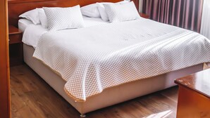 Uma cama bem arrumada com colcha branca e estampada, cabeceira de madeira e uma obra de arte pendurada na parede.