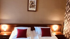 Quarto duplo com duas camas, cada uma com uma almofada e uma toalha dobrada. Há um quadro na parede e uma cortina estampada.