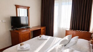 Quarto de hotel com cama, televisão, escrivaninha e janela com cortinas.
