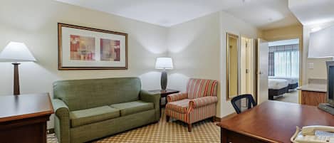 A hotel room with a green sofa, striped armchair, wooden desk, and a bed with a window view.