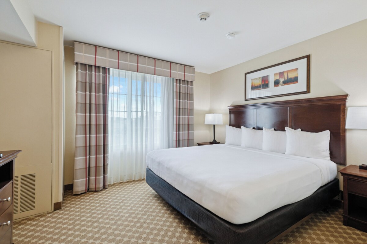 A hotel room with a large bed, a wooden headboard, a nightstand, a lamp, a wardrobe, and a window with curtains.