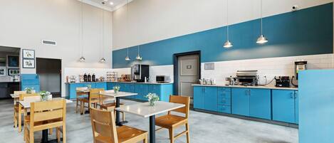 Een moderne keuken met blauwe kastjes, witte aanrechtbladen en hanglampen. Er staan houten tafels en stoelen, en een koelkast.