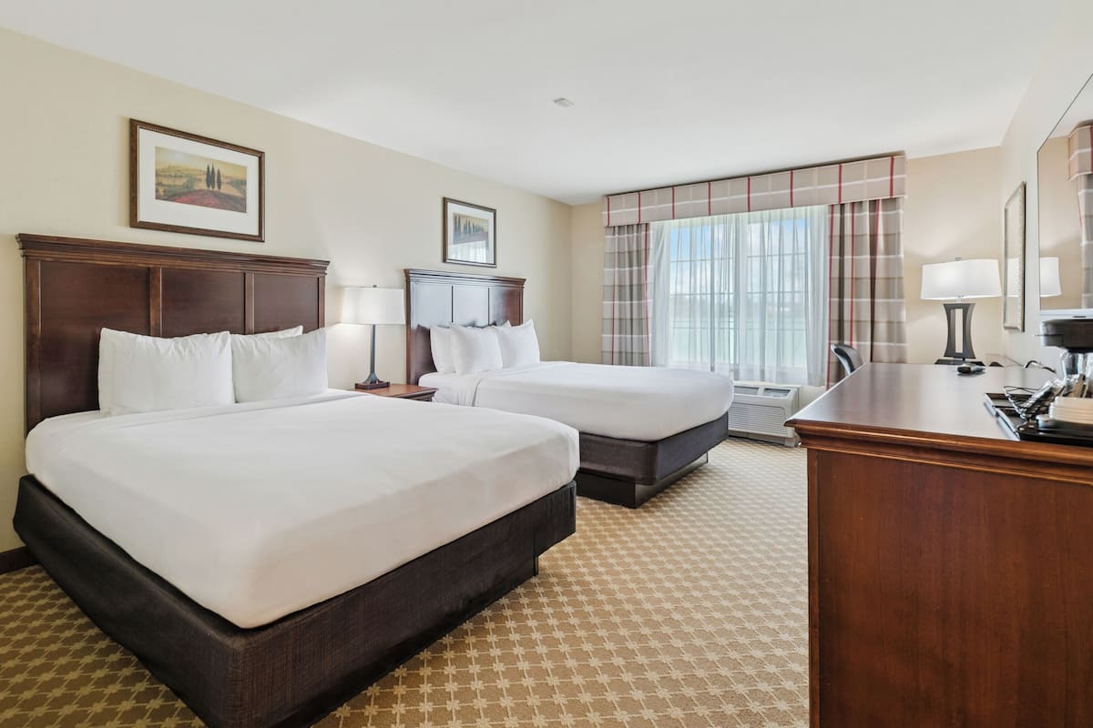 A hotel room with two beds, a desk, a chair, a lamp, and a large window with curtains.