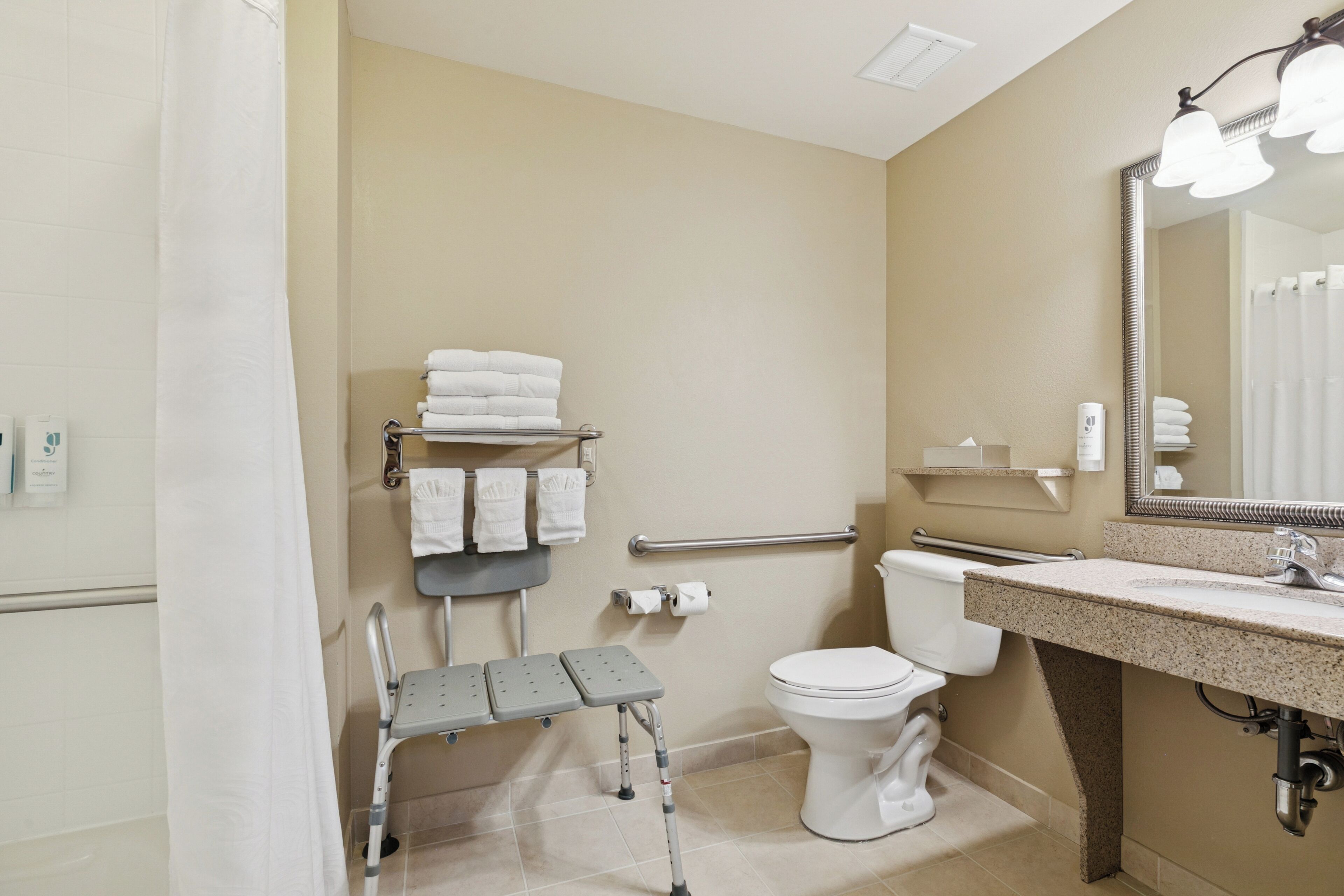 Baño con inodoro, lavamanos, espejo y un banco accesible para silla de ruedas.