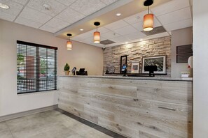 A hotel reception area with a wooden reception desk, a stone wall, and a window with blinds. - Country Inn & Suites by Radisson, Covington, LA (Covington)