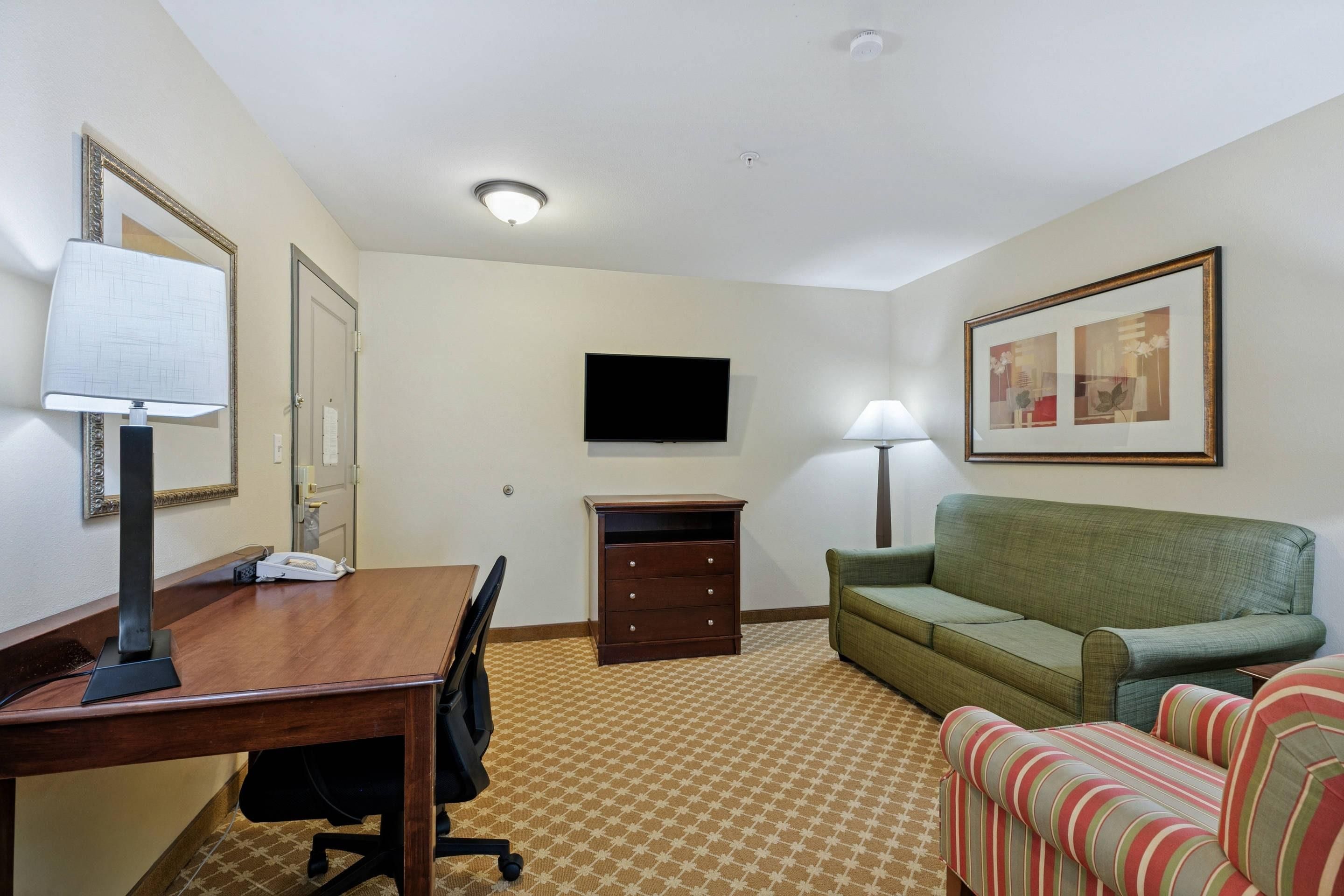 A hotel room with a wooden desk, a green sofa, a striped armchair, a flat-screen TV mounted on the wall, and a patterned carpet.
