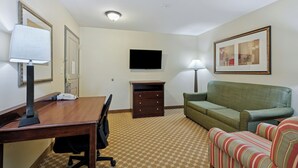 A hotel room with a wooden desk, a green sofa, a striped armchair, a flat-screen TV, a dresser, and a patterned carpet. - Country Inn & Suites by Radisson, Covington, LA (Covington)