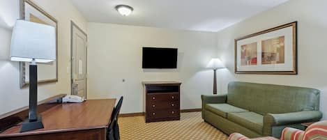 A hotel room with a wooden desk, a green sofa, a striped armchair, a flat-screen TV, a dresser, and a patterned carpet.