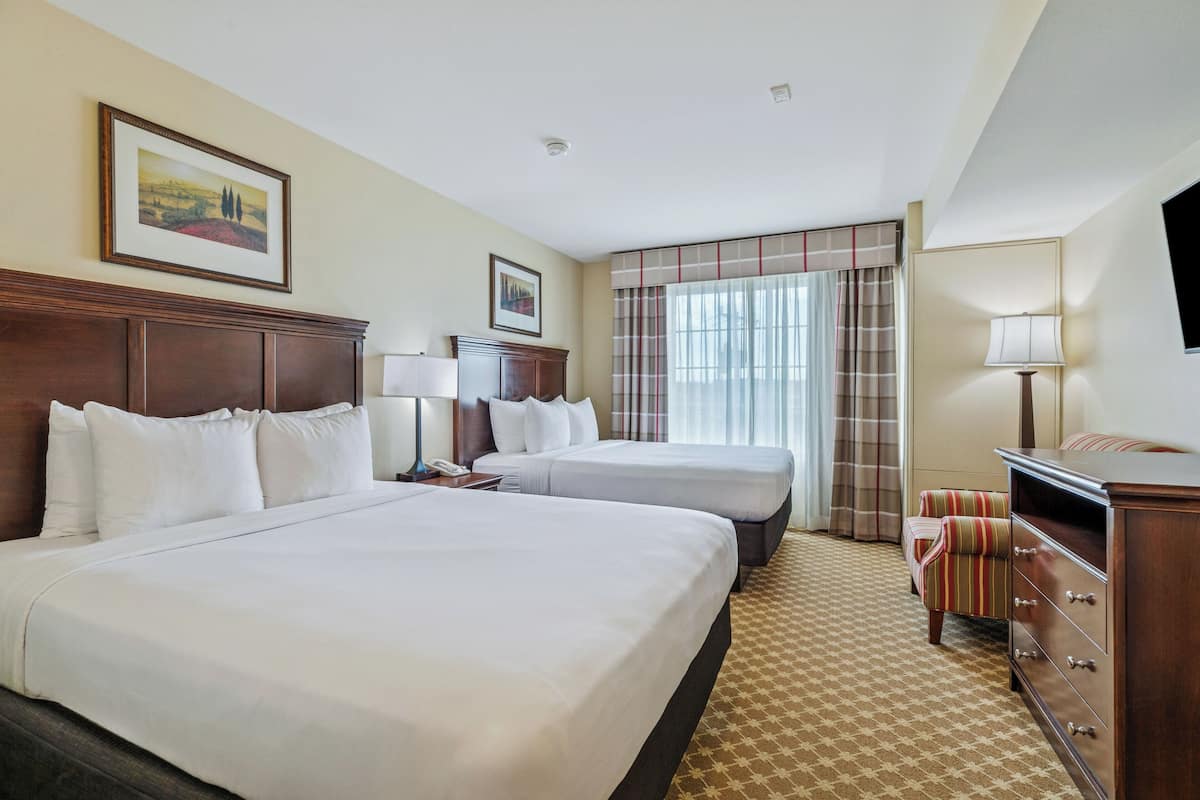 A hotel room with two large beds, a wooden dresser, a desk with a lamp, a chair, and a television.