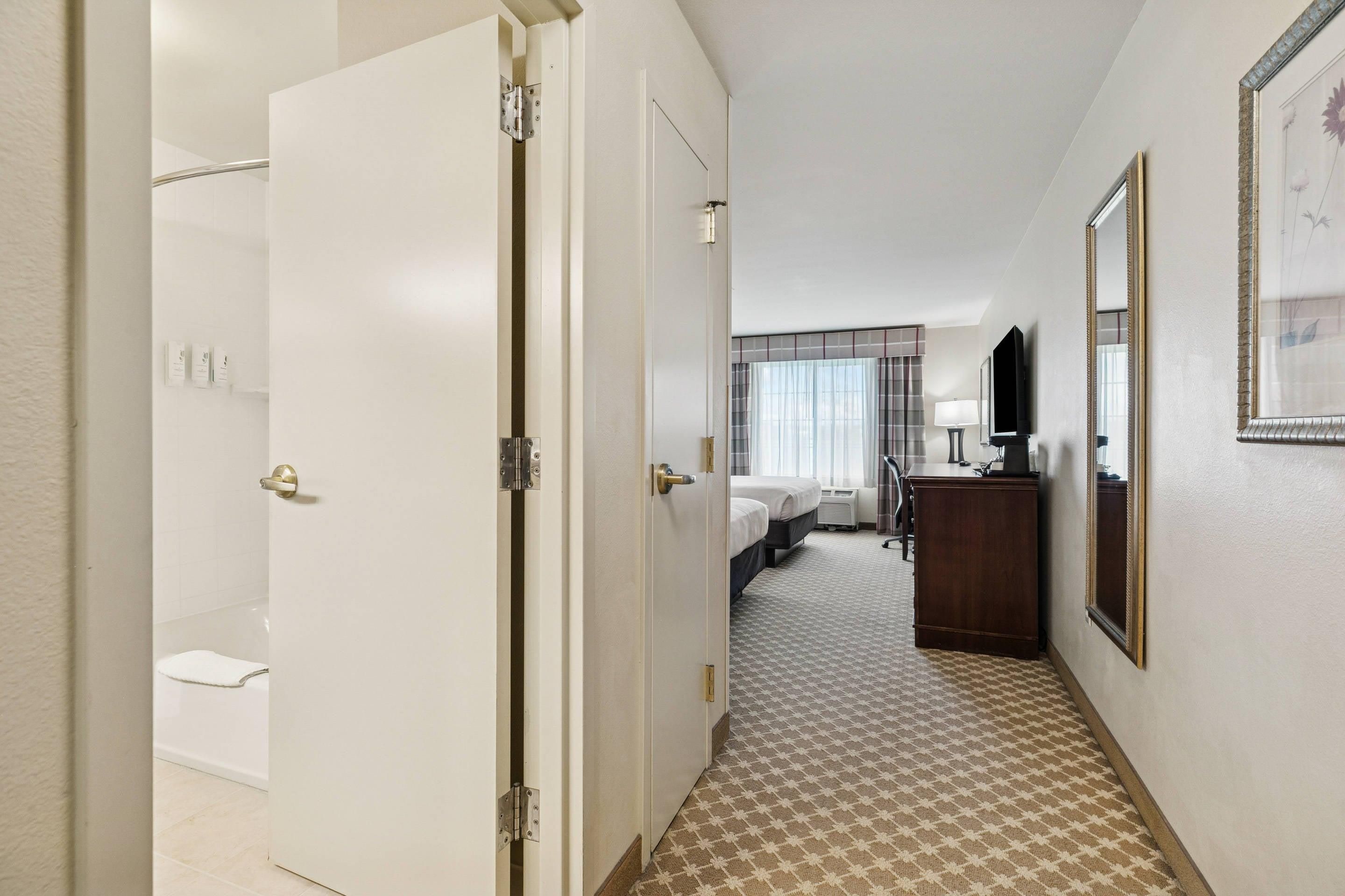A hotel hallway with a bathroom, a bed, a desk, and a mirror.