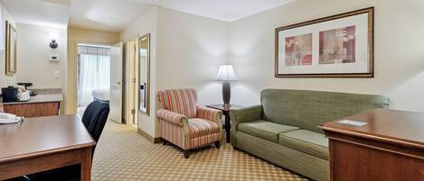 A hotel room with a desk, a striped armchair, a green sofa, a lamp, a framed picture, and a patterned carpet.