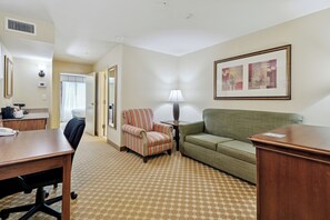 A hotel room with a wooden desk, a striped armchair, a green sofa, a lamp, and a framed picture on the wall.