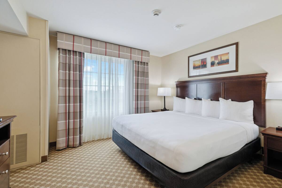 A hotel room with a large bed, a wooden headboard, a nightstand, a lamp, a wardrobe, and a window with curtains.