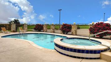 A swimming pool with lounge chairs and a hot tub, surrounded by a stone wall and a metal fence.