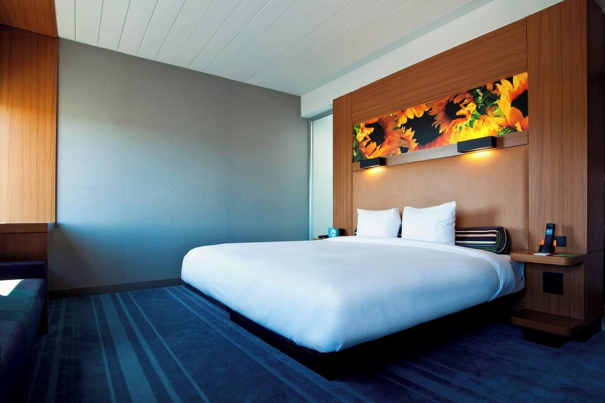 A hotel room with a large bed, a nightstand, a desk, and a wall-mounted artwork.