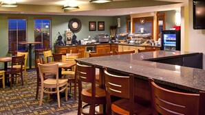 A hotel kitchen with a central island, wooden chairs, a coffee station, and a refrigerator. - AmericInn by Wyndham Anamosa (Anamosa)