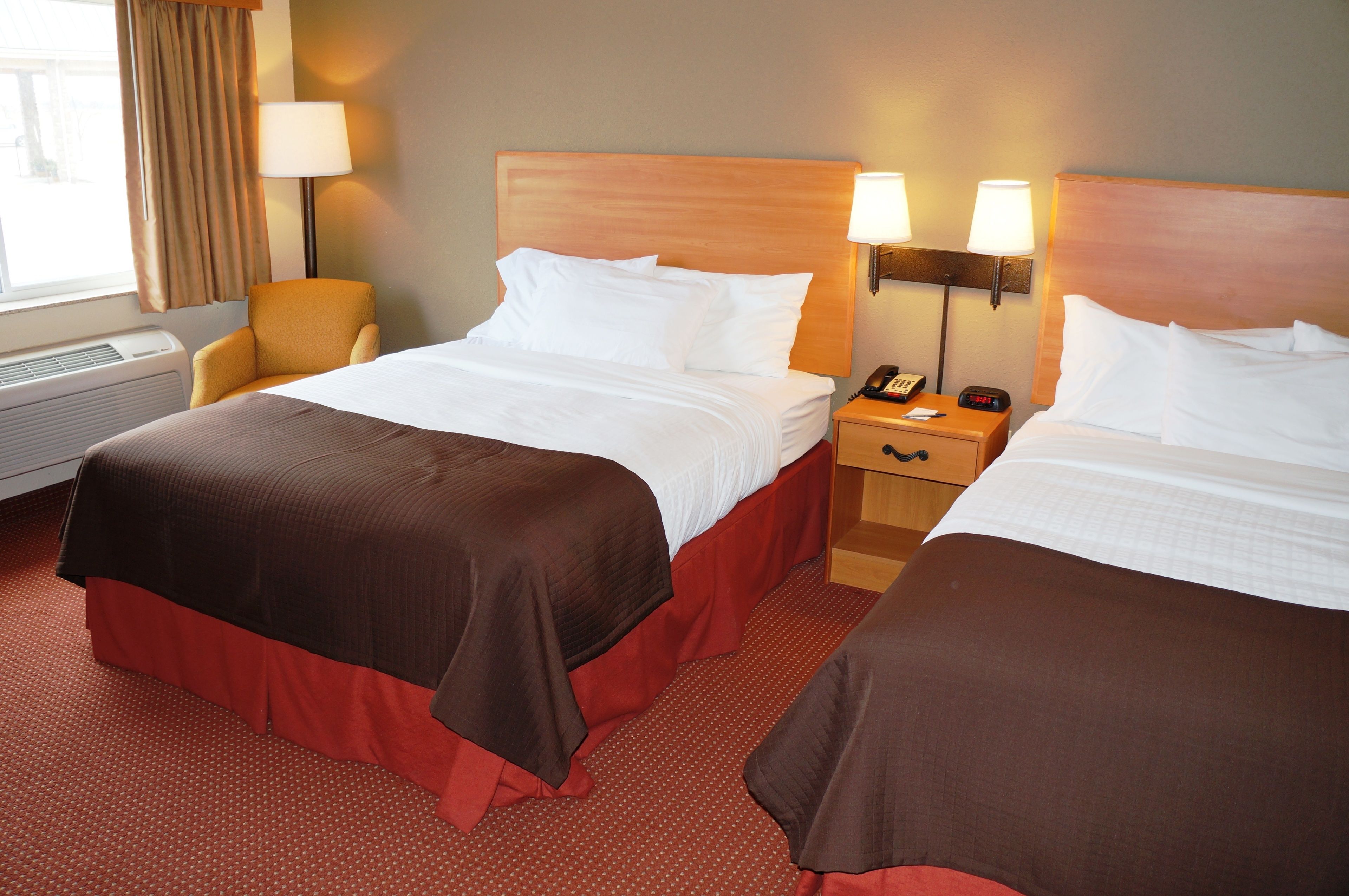 Quarto de hotel com duas camas, uma cadeira, mesa de cabeceira com abajur e janela com cortinas.