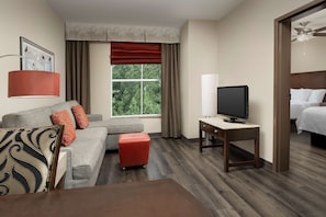 A hotel room with a grey sofa, red ottoman, a TV on a wooden table, and a bed with white linens.