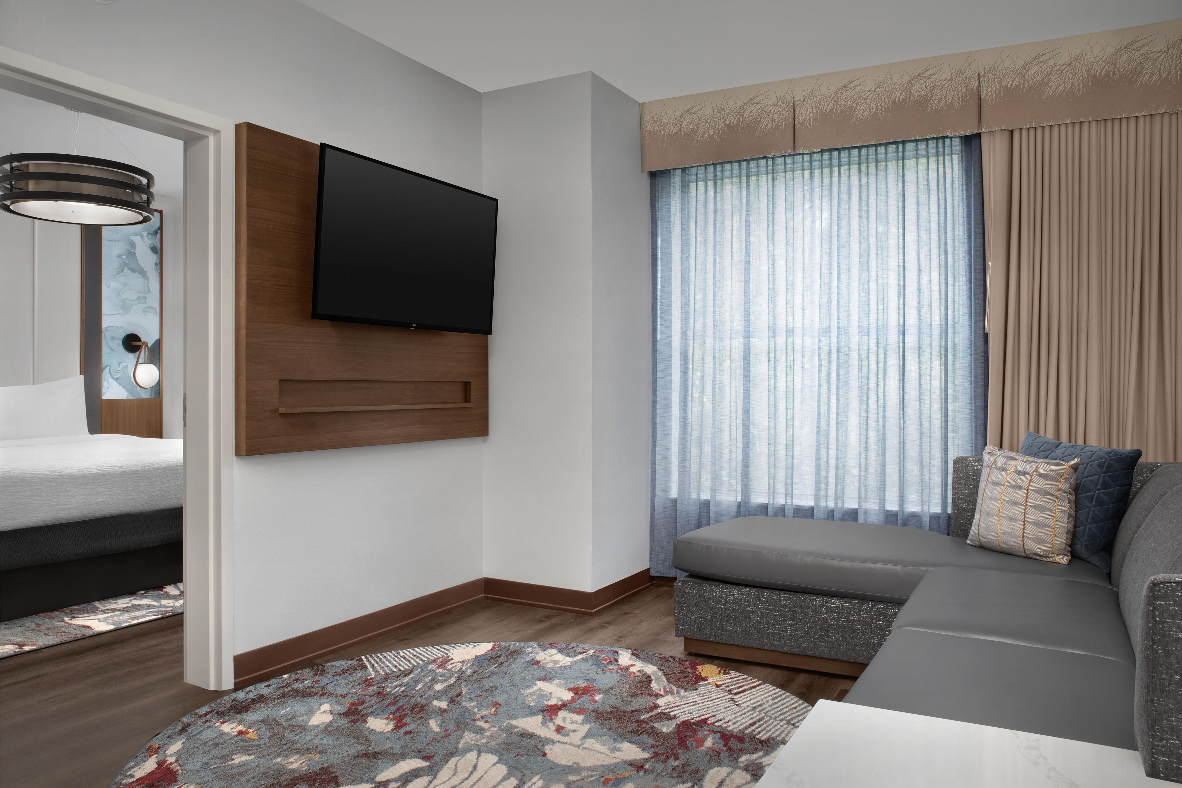 A modern living room with a flat-screen TV mounted on the wall, a sofa, a patterned rug, and a window with sheer curtains.