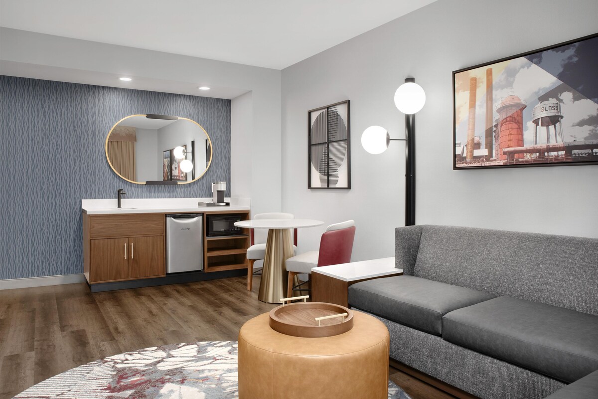 A modern living room with a grey sofa, a round wooden coffee table, a white round table, a red chair, a kitchen area with a sink and cabinets, and a wall with a patterned wallpaper and artwork.