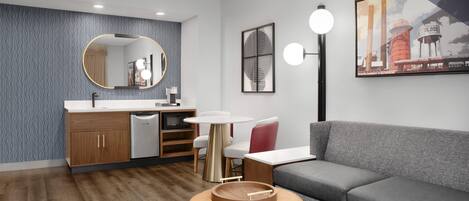 Una sala moderna con un sofá gris, una mesa de centro redonda de madera, una mesa redonda blanca, una silla roja, una zona de cocina con un fregadero y armarios, y una pared con papel tapiz estampado y obras de arte.