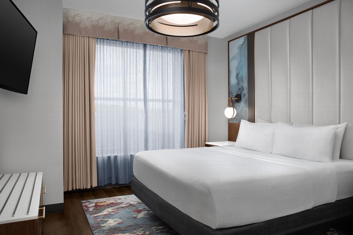A hotel room with a large bed, a TV, a painting on the wall, and a window with curtains.