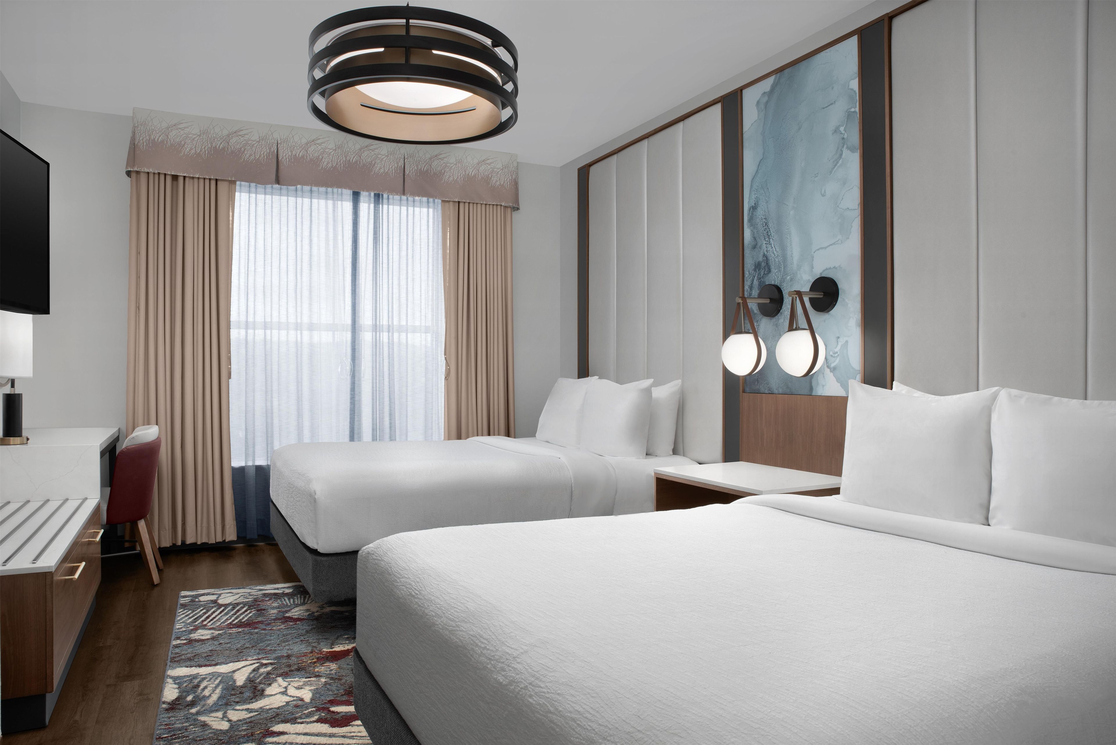A hotel room with two beds, a desk, a chair, a TV, and a large window with curtains.