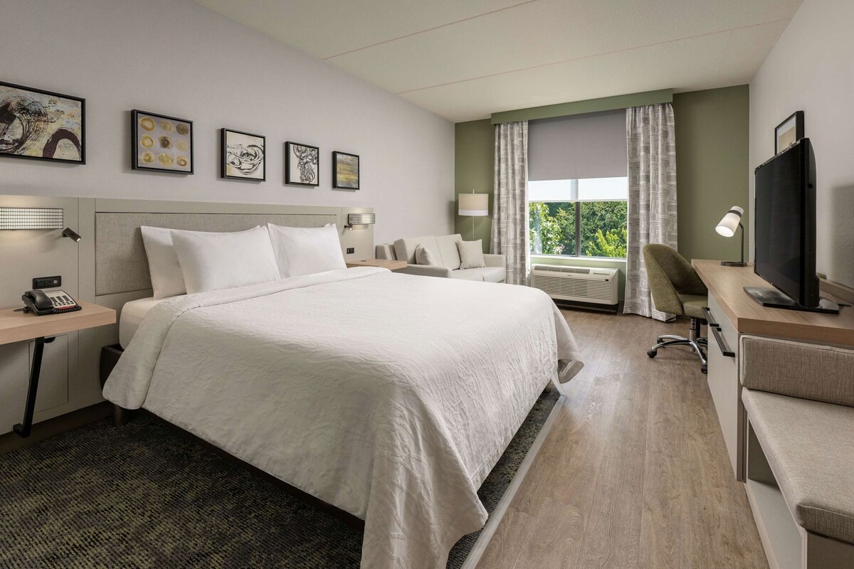 A hotel room with a large bed, a desk with a computer, a chair, a TV, and artwork on the wall.