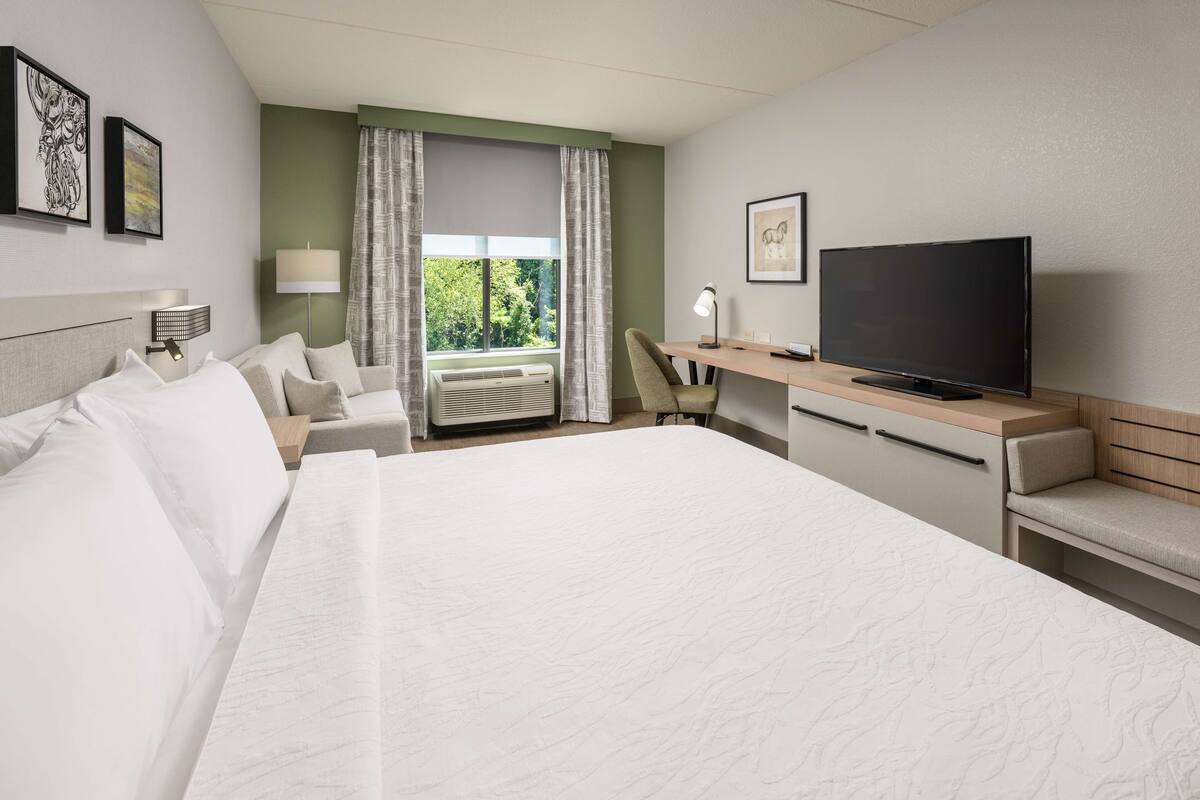 A hotel room with a large bed, a desk with a flat-screen TV, a chair, and a window with curtains.