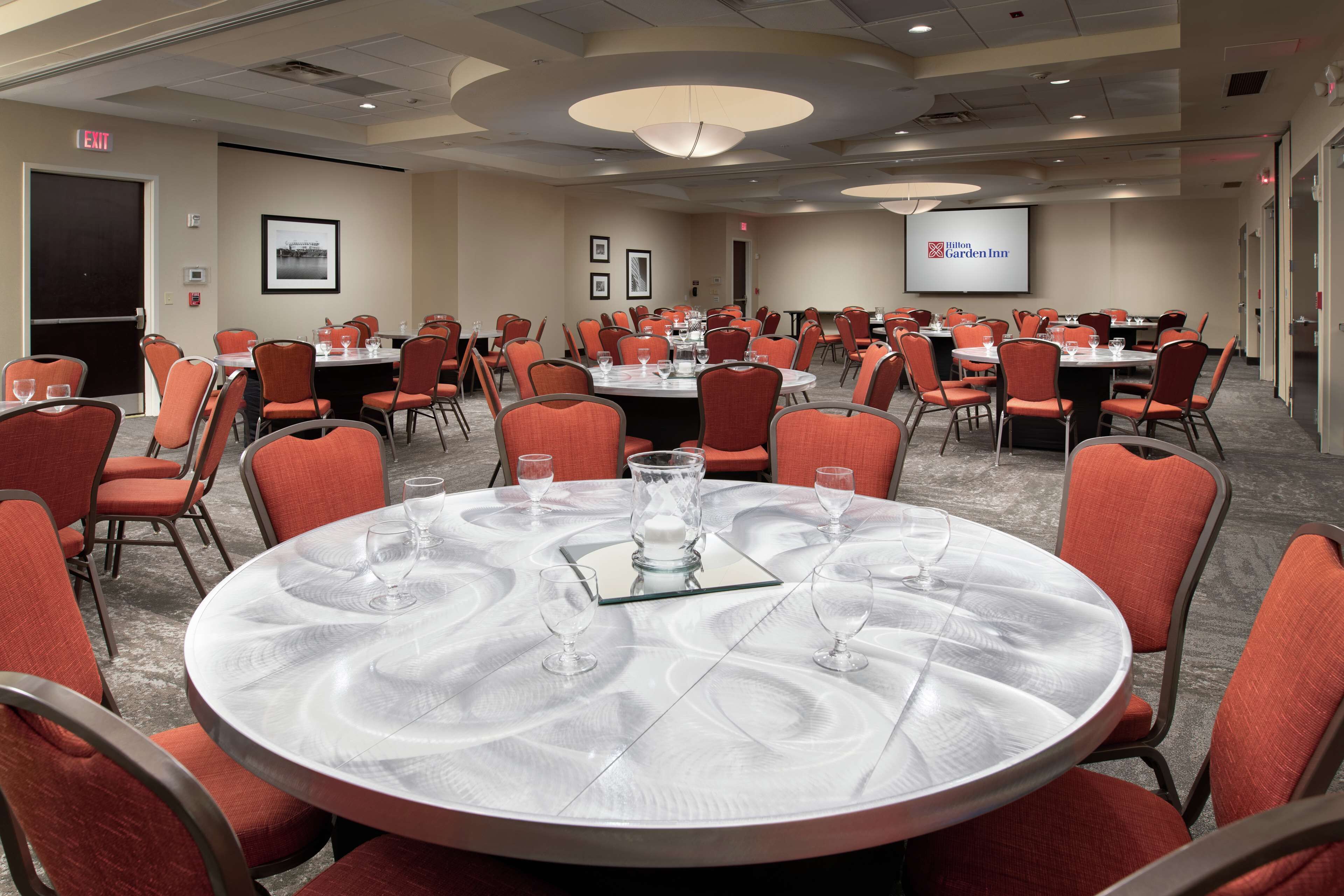 A conference room with multiple tables and chairs, a projector screen displaying the Hilton Garden Inn logo, and framed pictures on the wall.