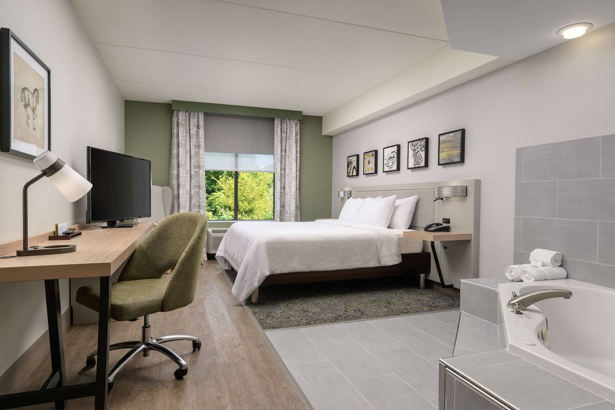 A hotel room with a bed, desk, chair, TV, and a bathroom with a bathtub.