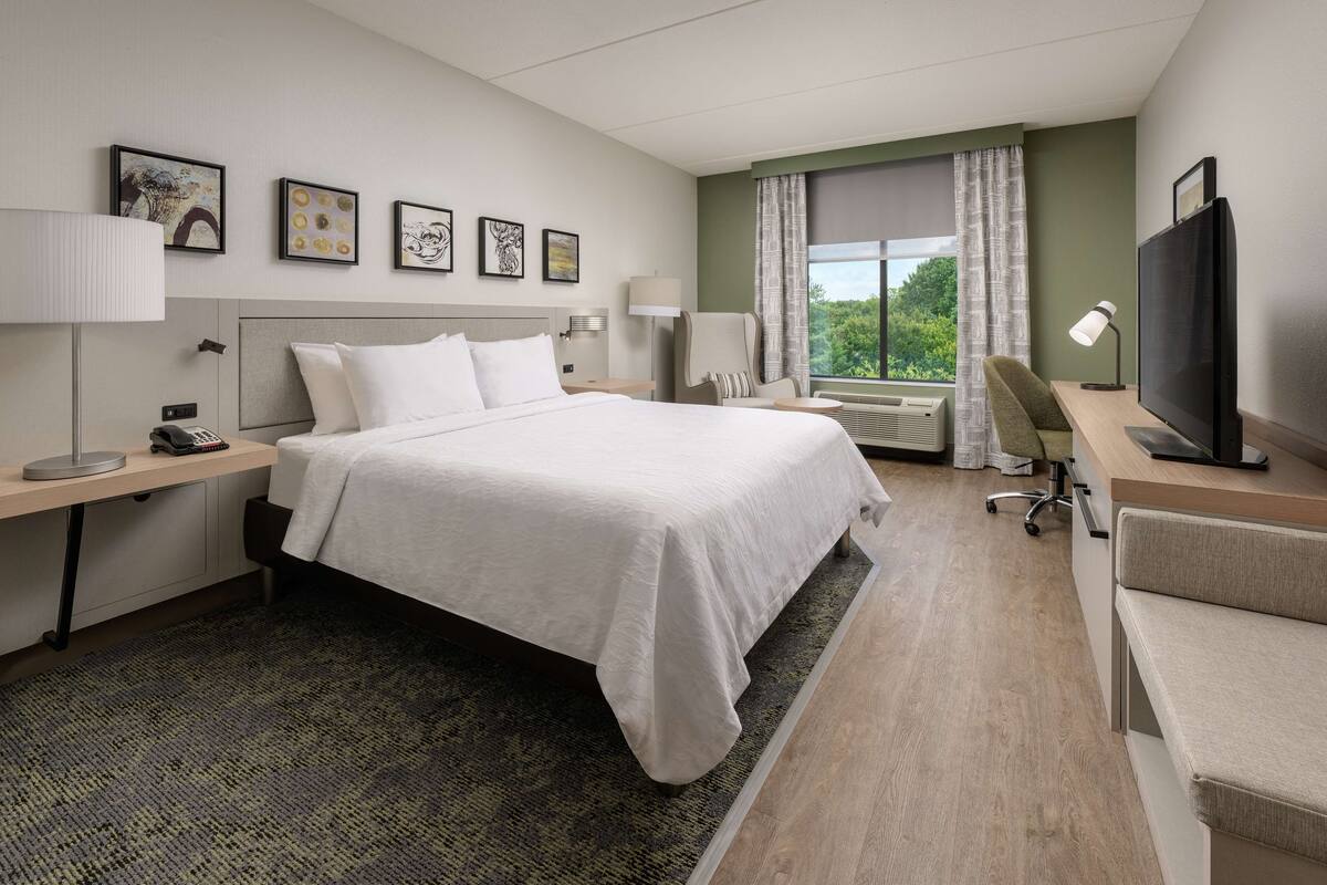 A hotel room with a large bed, a desk, a chair, a TV, and a window with curtains.