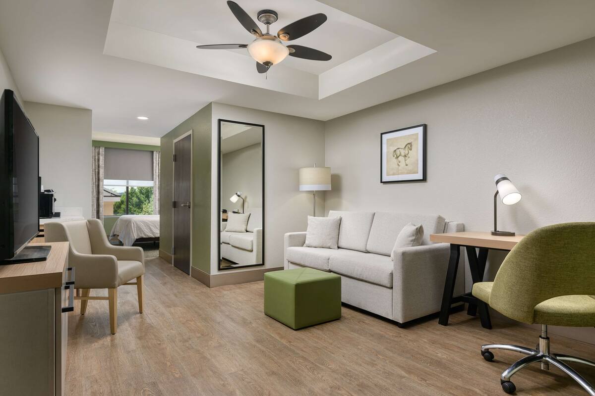 A modern hotel room with a wooden floor, a ceiling fan, a sofa, a desk, a chair, and a green ottoman.