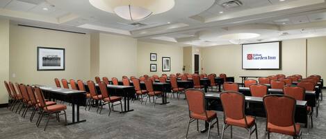 A conference room with rows of tables and chairs, a projector screen displaying the Hilton Garden Inn logo, and framed pictures on the walls.