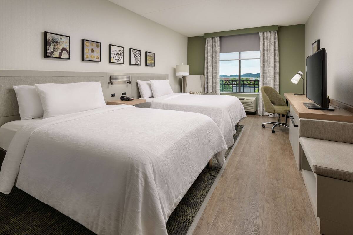 A hotel room with two beds, a desk, a chair, a TV, and a window with a view.