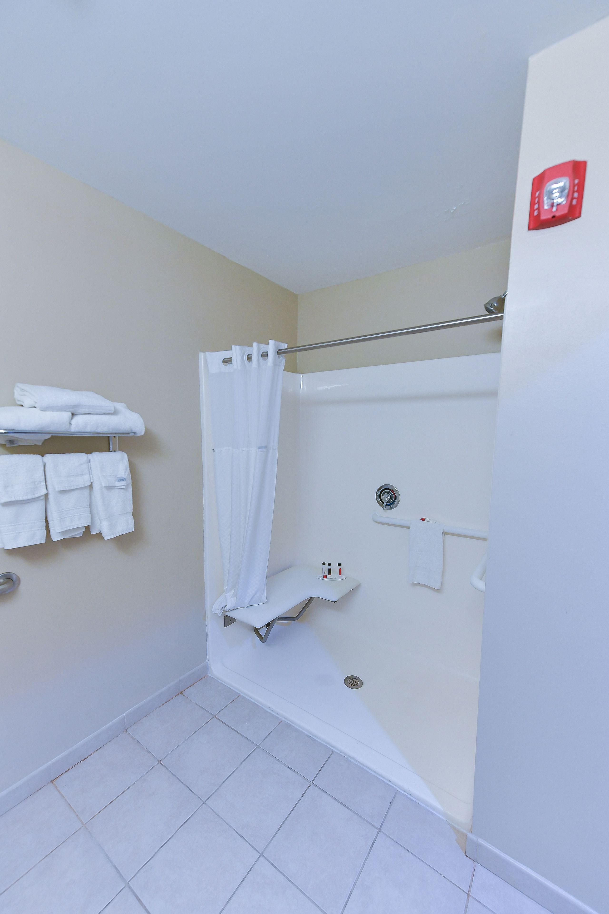 Een badkamer met een douche, een handdoekrek met opgevouwen handdoeken en een veiligheidslamp aan de muur.