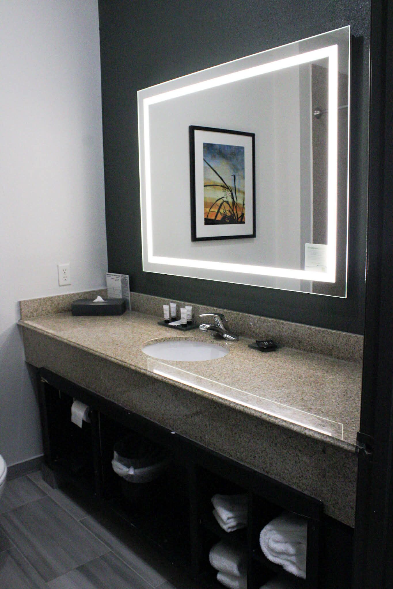 Un baño moderno con encimera de granito, un lavabo blanco, una obra de arte enmarcadasobre el espejo y un perchero debajo.