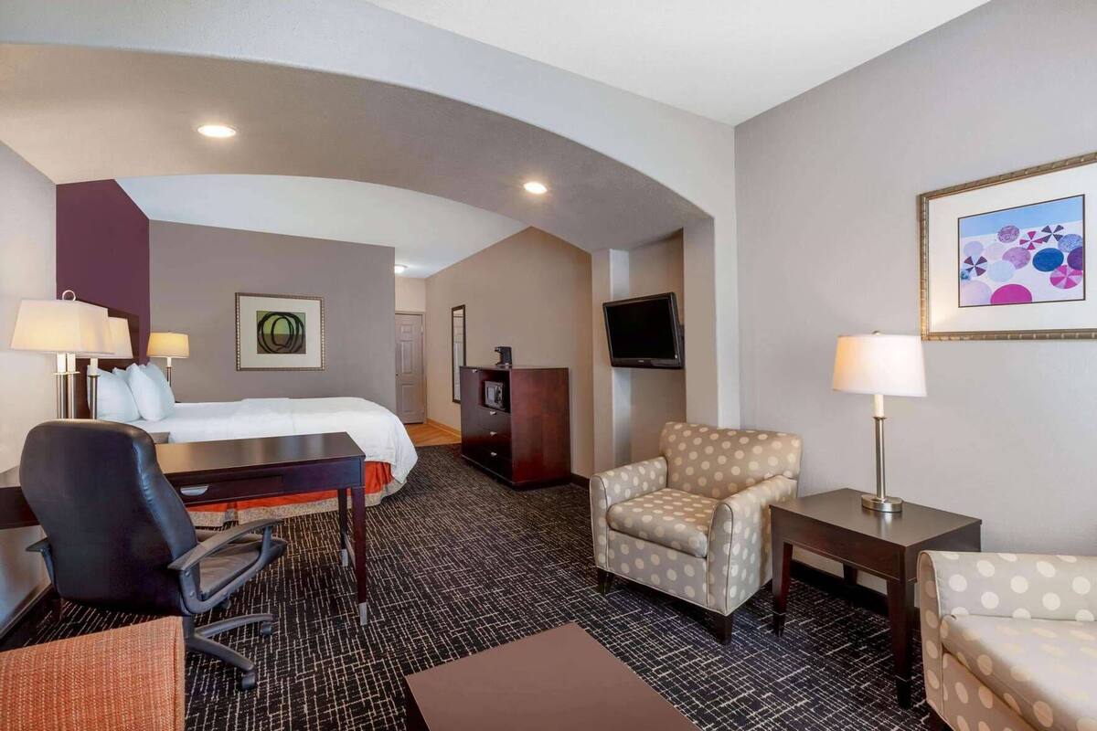 A hotel room with a bed, a desk, a chair, a TV, and a painting on the wall.