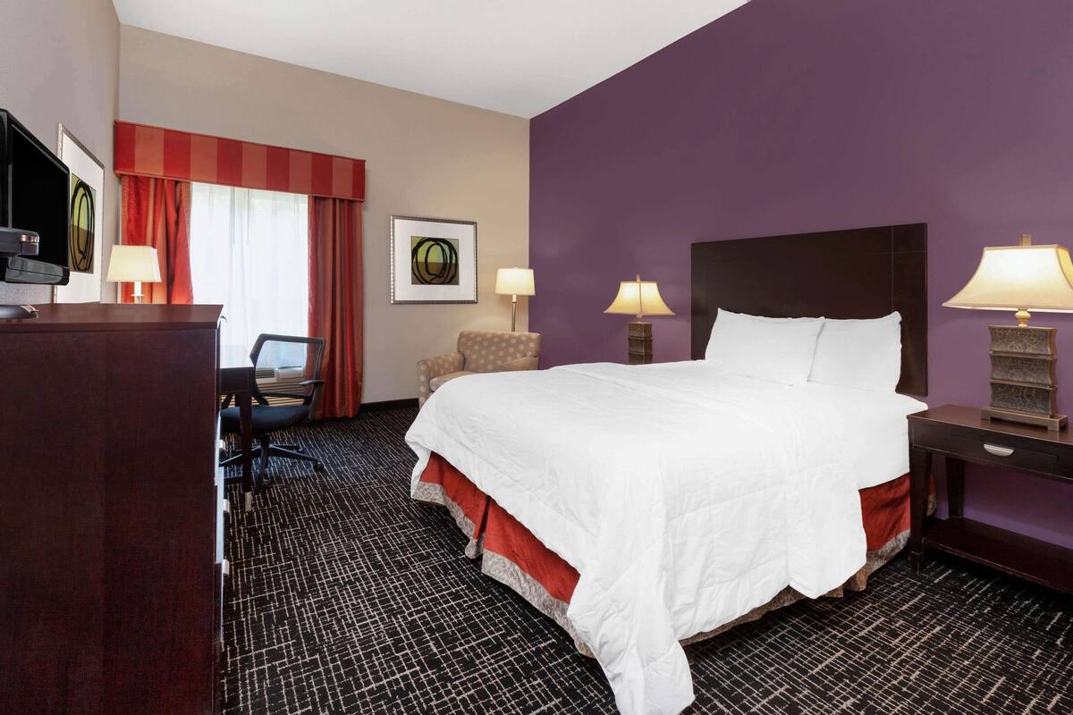 A hotel room with a large bed, a desk with a computer, a chair, two lamps, and a television.